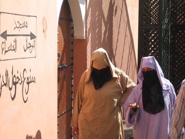 Two veiled women walk past a pink wall with Arabic signs