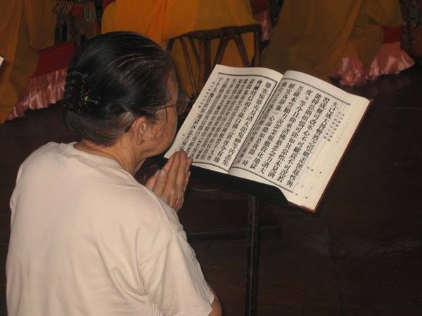 Elderly woman praying before an open Chinese sutra book in a temple