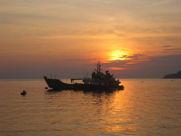 Ferry silhouetted on golden sea, nearby canoes, hill on the right