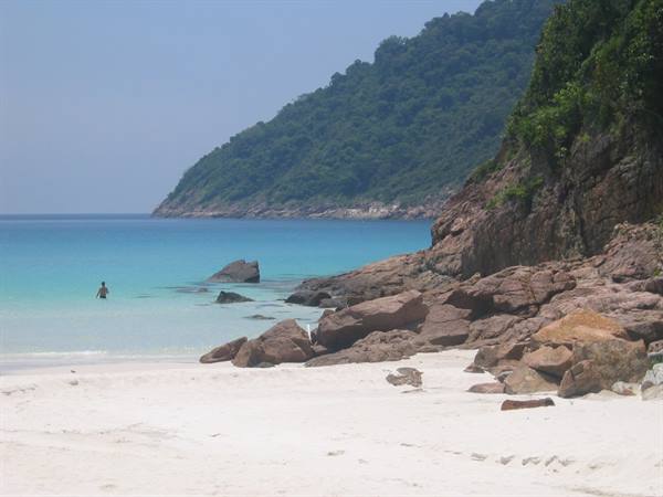 Swimmer in turquoise water among red rocks, white sand, green hill