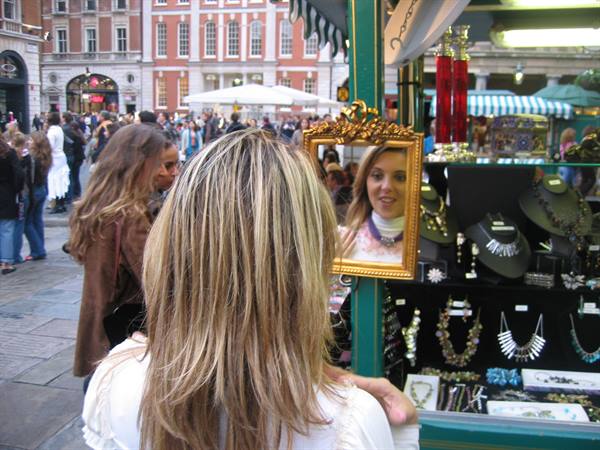 Blonde woman mirrors herself in a gilded frame at a jewelry stall, crowded square
