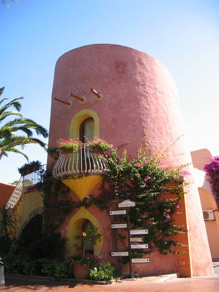 Pink stucco round tower with white balcony, flowering bougainvillea and signs, summer sun