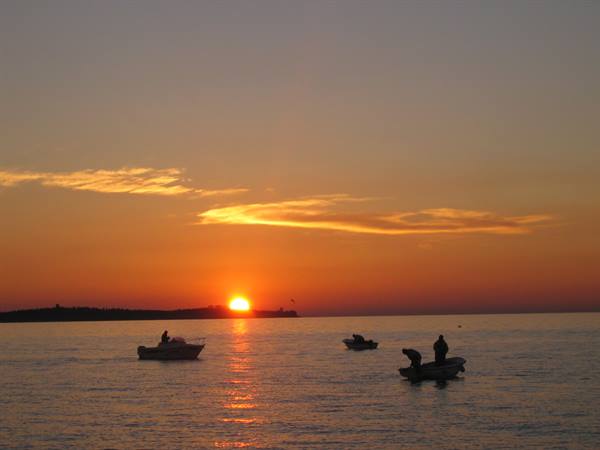 Fishermen on small boats offshore, low sun glows orange over sea and sky