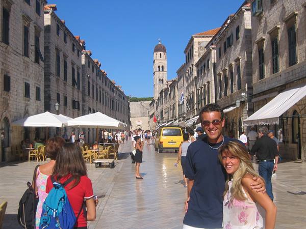 Smiling tourists on Stradun, Dubrovnik, with cafés, umbrellas and bell tower