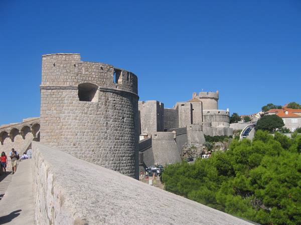 Tourists walk along Dubrovnik's stone walls with fortified towers under sunny sky