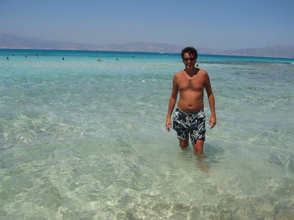 Man in dark swim trunks walks through clear turquoise water, swimmers nearby