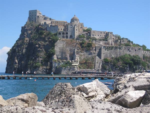 Aragonese Castle on volcanic rock in Ischia, tourists on pier among pale boulders