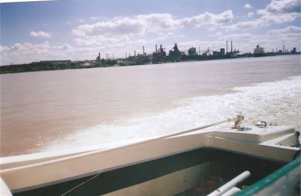 Boat wake on muddy river, industrial plants along the shore under clouds