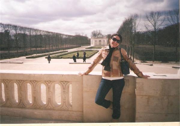 Woman in sunglasses leaning on carved stone balustrade, tree-lined avenue and white pavilion, Parc de Sceaux