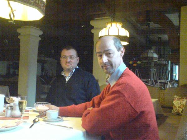 Two men seated at a set table, glowing chandeliers, columns in the hall
