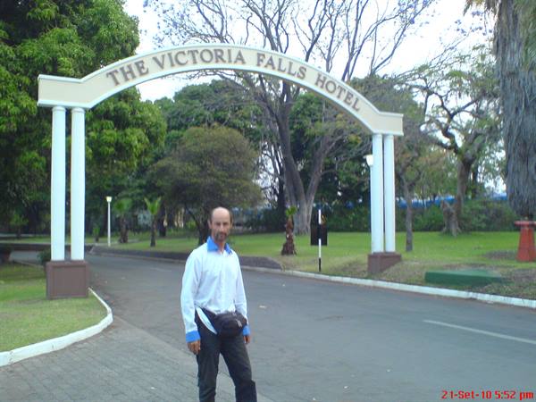 Man under the white entrance arch of the Victoria Falls Hotel, road and green gardens