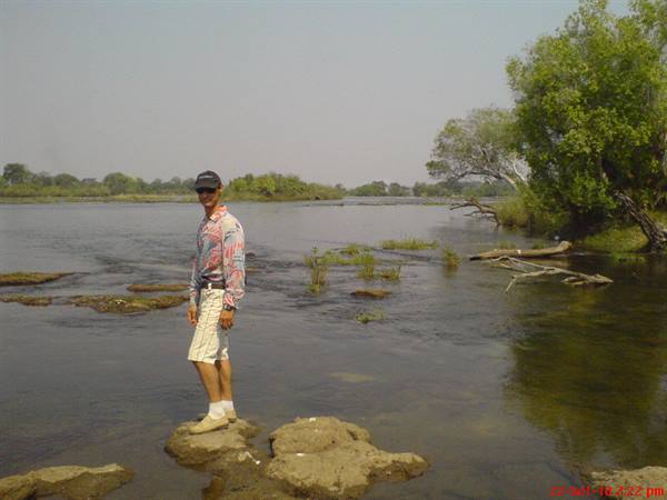 Tourist in floral shirt standing on a rock in the Zambezi river, shallow water and green trees