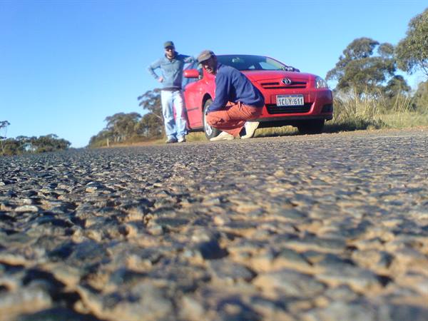 Two men by a red Toyota, one crouching beside the front wheel
