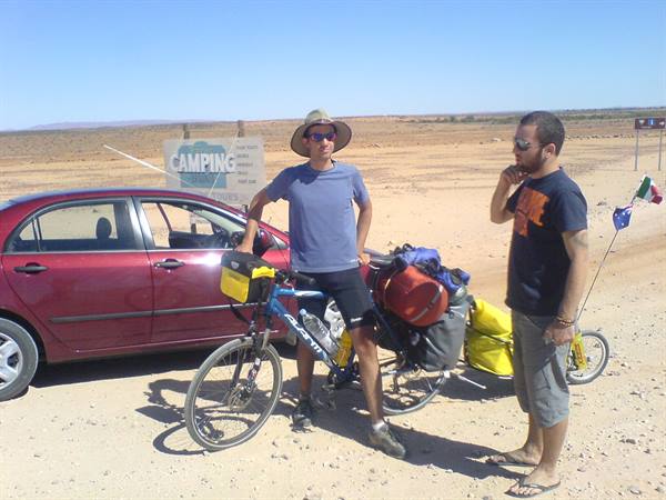 Two cyclists with loaded touring bike beside a red car in the desert near camping sign