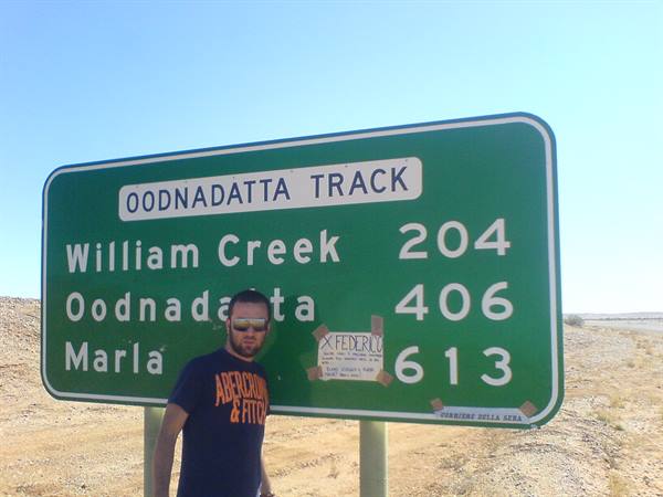 Man in sunglasses beside the green Oodnadatta Track road sign in the desert