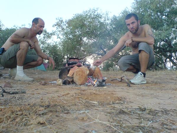 Two men crouch lighting a stone campfire on red dirt among trees