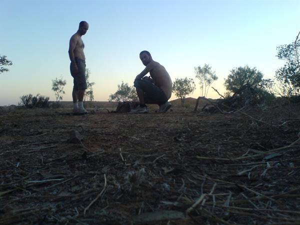 Two men crouching in sandy scrubland at dusk, warm light