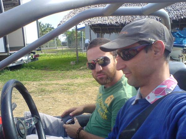 Two men in sunglasses riding a buggy in front of thatched huts