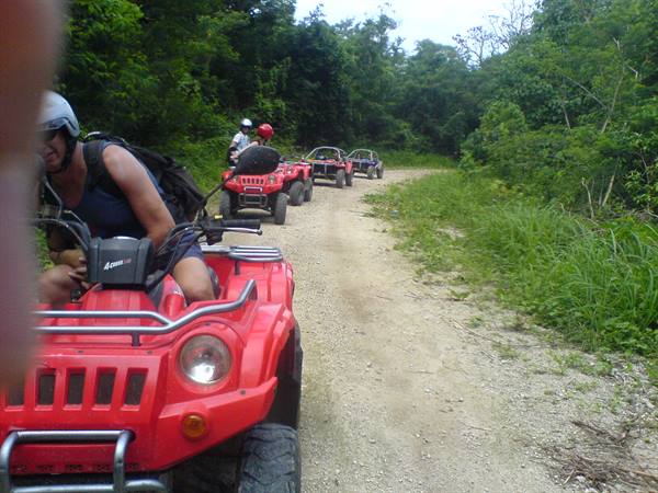 Helmeted tourists riding red ATVs on dirt road through tropical greenery