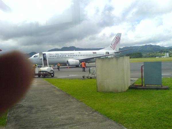 Air Vanuatu plane parked on tarmac, workers boarding, green hills under cloudy sky
