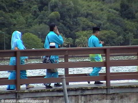 People in blue outfits and light veil walk along a wooden footbridge above the lake