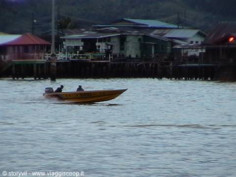 Yellow speedboat with two people rushes past wooden stilt houses on water