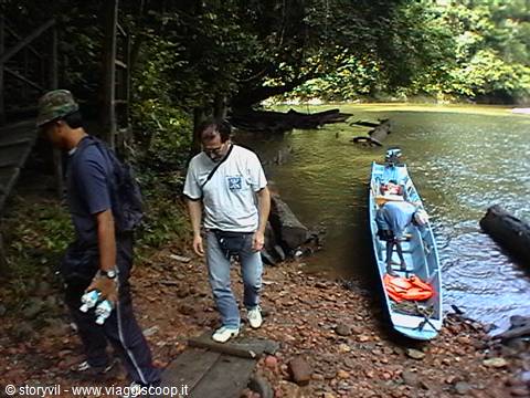 Tourists walk down rocky shore beside a long blue boat on river