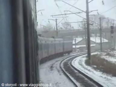 Train on snowy tracks seen through window, catenary poles and falling snow