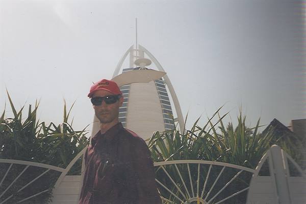 Man in red cap and sunglasses in front of Burj Al Arab, palms and white fence
