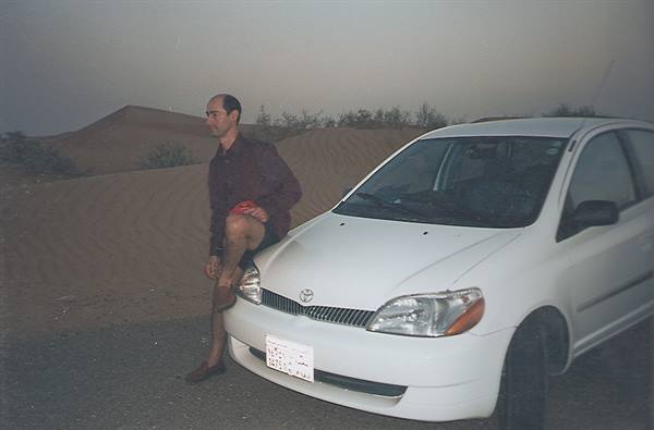 Man beside a white Toyota in front of desert sand dunes at dusk