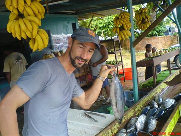 Man in cap holding a fish at market stall among hanging bananas
