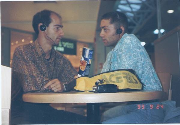 Two men wearing headphones chat at a cafe table with a yellow bag
