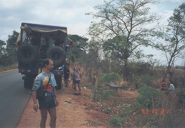 Travelers beside an open safari truck on a dirt road among dry trees