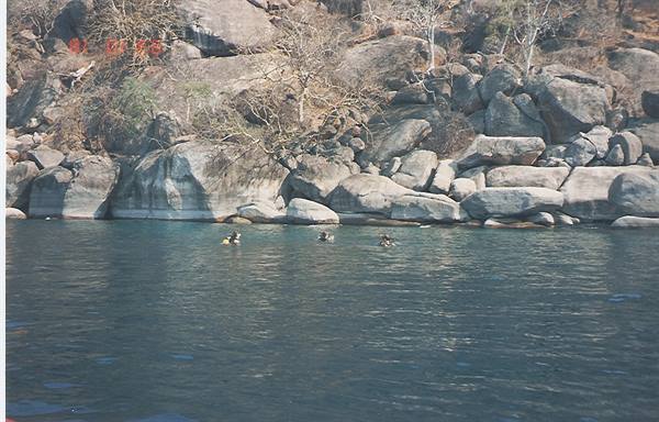 Swimmers in dark green water in front of granite boulders and bare trees