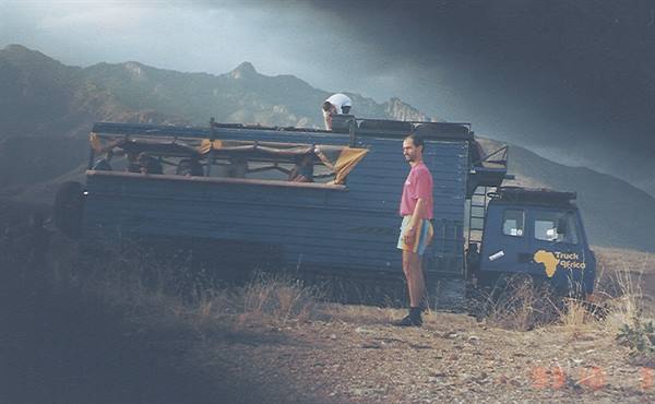 Man in pink shirt beside blue overland truck among arid mountains