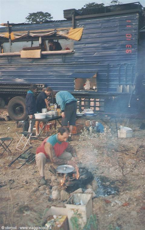 Blue overland truck at campsite, man crouching lighting fire, women preparing dinner