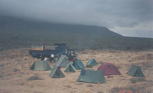 Tents and a truck camped on dry scrubland below a cloud-covered hill