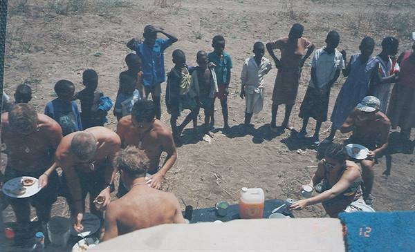 African children watch tourists eating outdoors on sandy ground