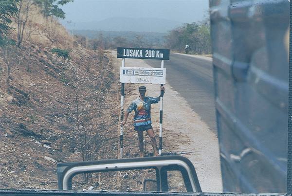 Man leaning on the Lusaka 200 km road sign by the asphalt