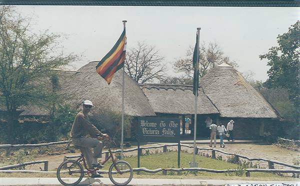 Cyclist passing thatched cottages at Victoria Falls entrance, Zimbabwean flag flying
