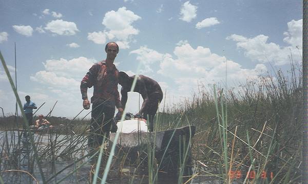 Two men in a dugout canoe among reeds, white cargo, cloudy sky