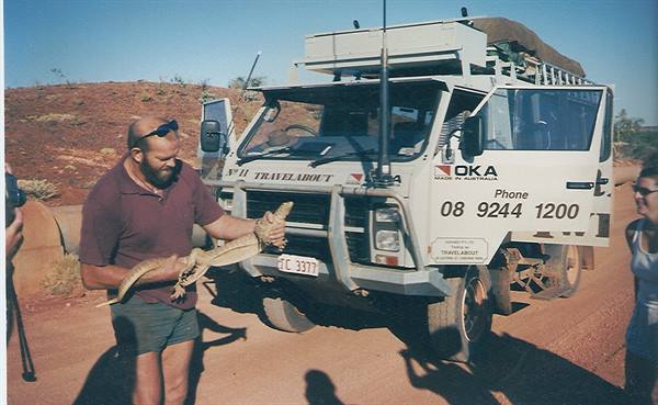 Bearded man holds a goanna beside a white 4x4 truck on red dirt road