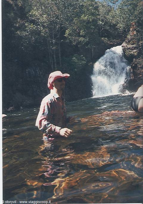Man in pink hat wading in water below a waterfall among rocks and trees