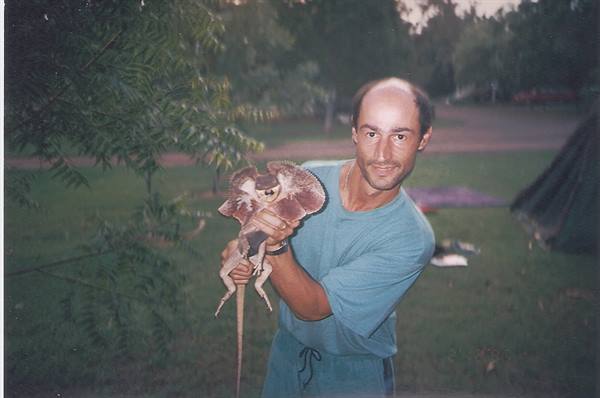 Man in blue t-shirt holds a frilled lizard at a leafy campsite