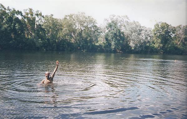Man swimming in river with arm raised, tree-lined banks behind