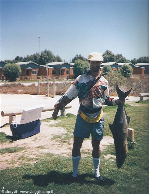 Man in yellow hat holding a caught shark near wooden cabins