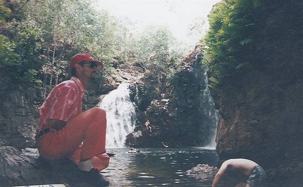 Man in red crouching beside waterfalls among rocks and ferns