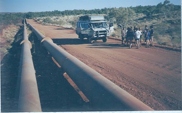 Tourist group beside a four-wheel-drive vehicle on red dirt road, metal pipeline along the edge