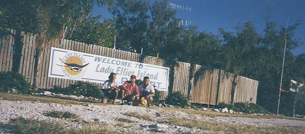 Three people crouch before the Lady Elliot Island sign, sand and palm trees