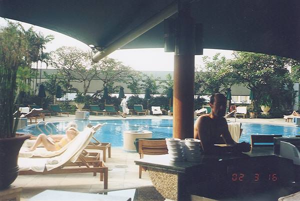 Man lounging by hotel pool under canopy, deck chairs and stacked cups nearby